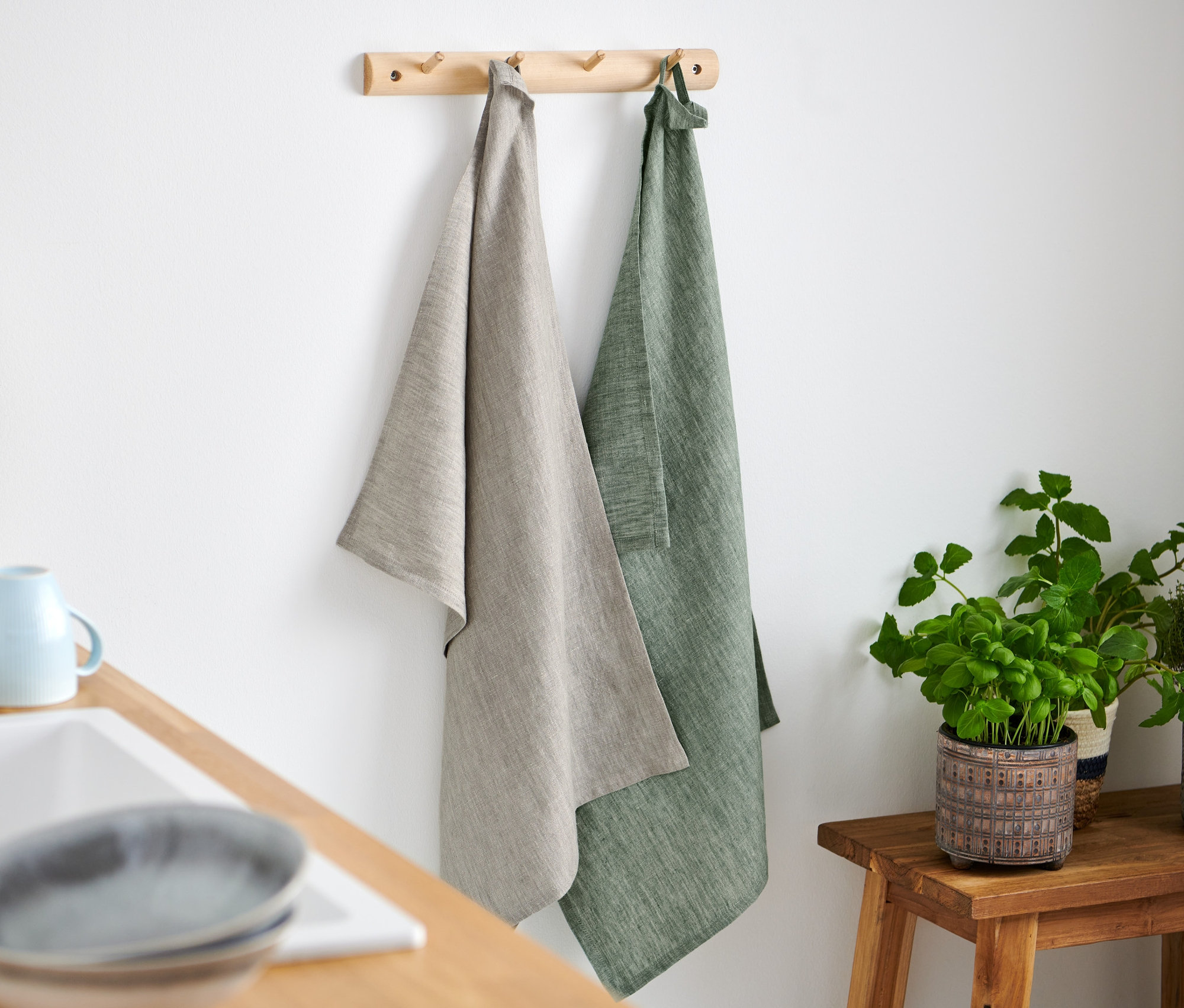 Dwie lniane ściereczki do naczyń wiszą na drewnianym wieszaku, jedna beżowa, druga zielona.
