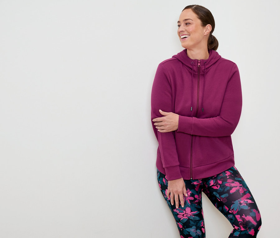 Kobieta w purpurowej bluzie z kapturem na zamek i kwiecistych legginsach opiera się o białą ścianę i uśmiecha się.