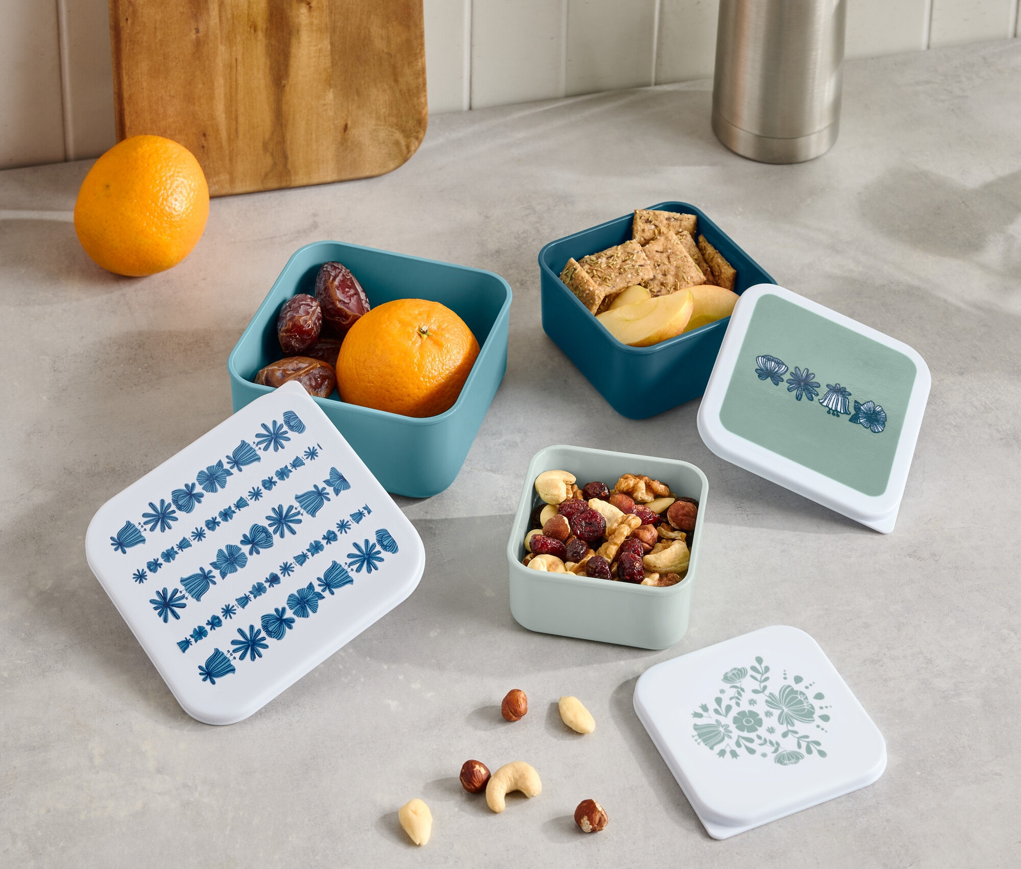 Na blacie stoją termos i drewniana deska do krojenia obok czterech małych otwartych pudełek z owocami, krakersami i orzechami oraz czterech pokrywek z różnymi wzorami.
