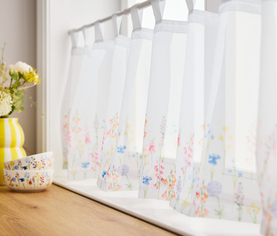 Kwiaty w żółtym wazonie i dwie miski na drewnianej desce przed oknami z zasłonami w kwiaty.