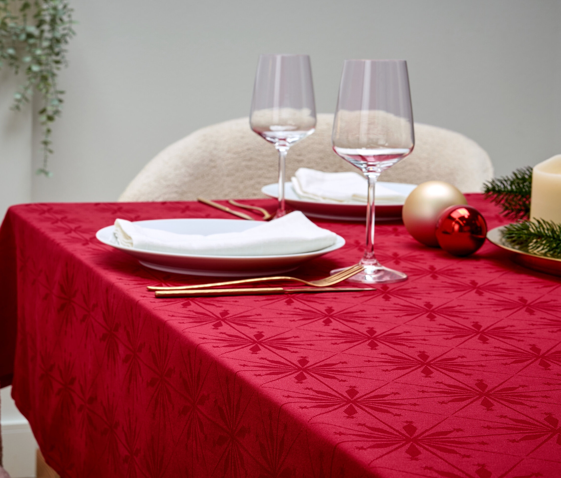Stół nakryty czerwonym obrusem, białymi talerzami, kieliszkami do wina, złotymi sztućcami, ozdobami i świecą.