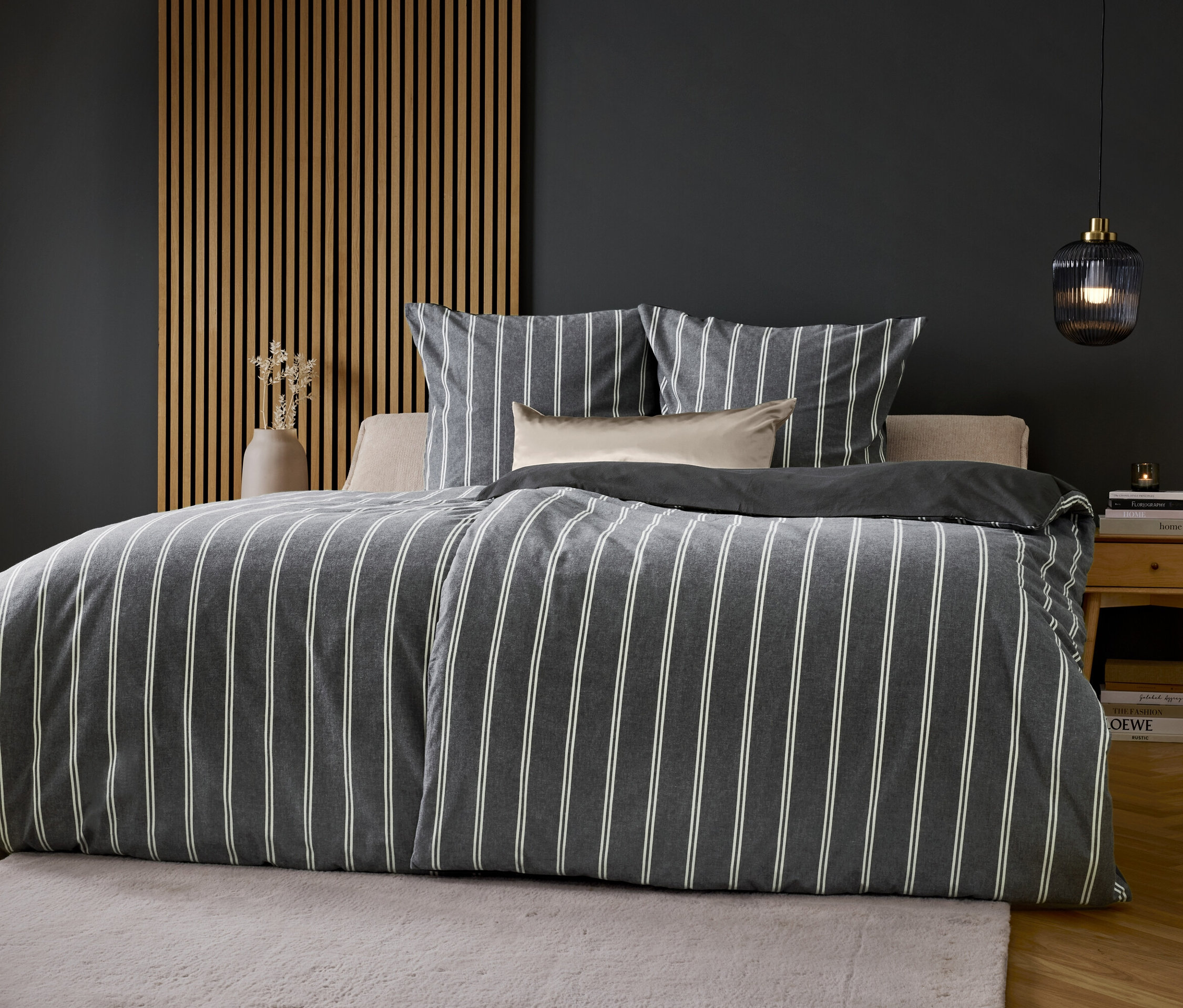 Łóżko z szaro-białą pościelą w paski na tle szarej ściany.
