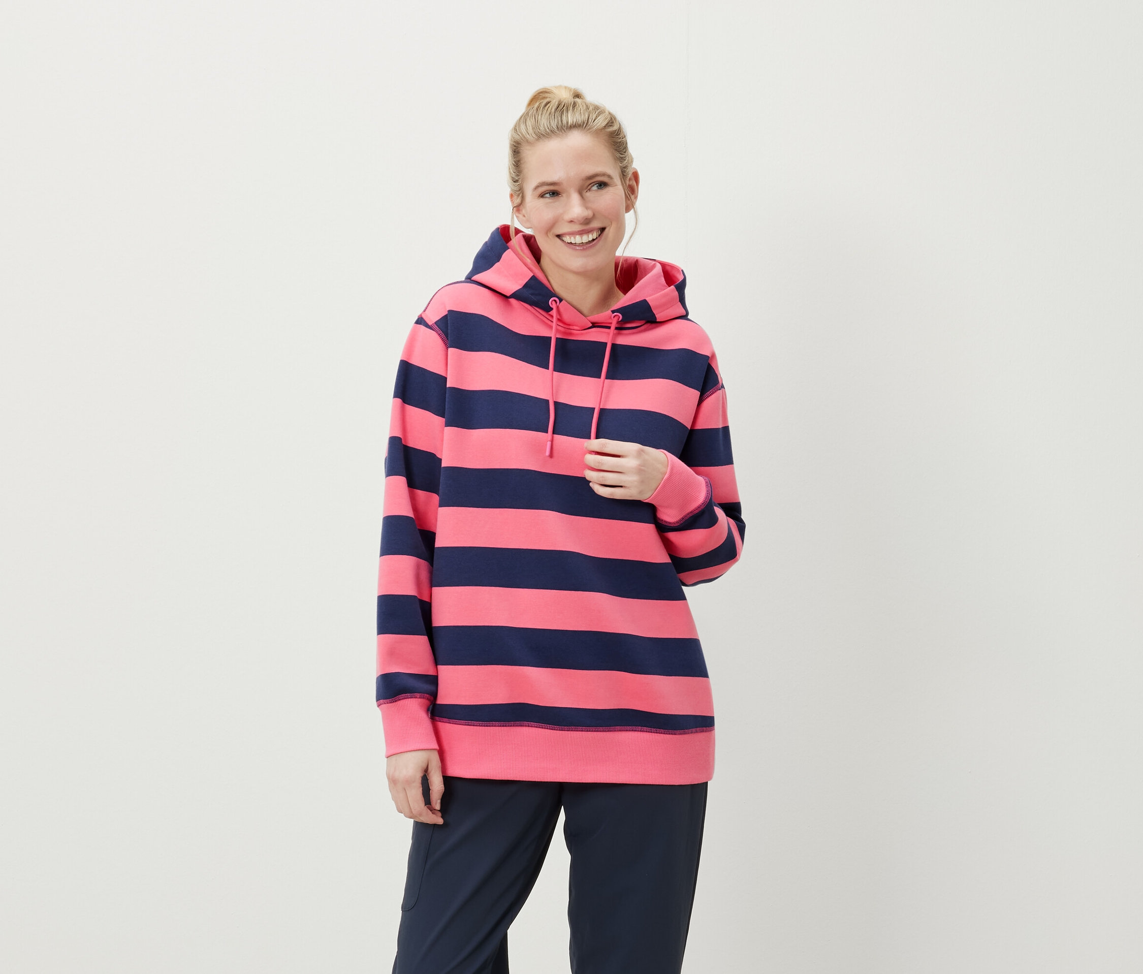 Uśmiechnięta blondynka w różowo-niebieskiej bluzie z kapturem w paski i granatowych spodniach.