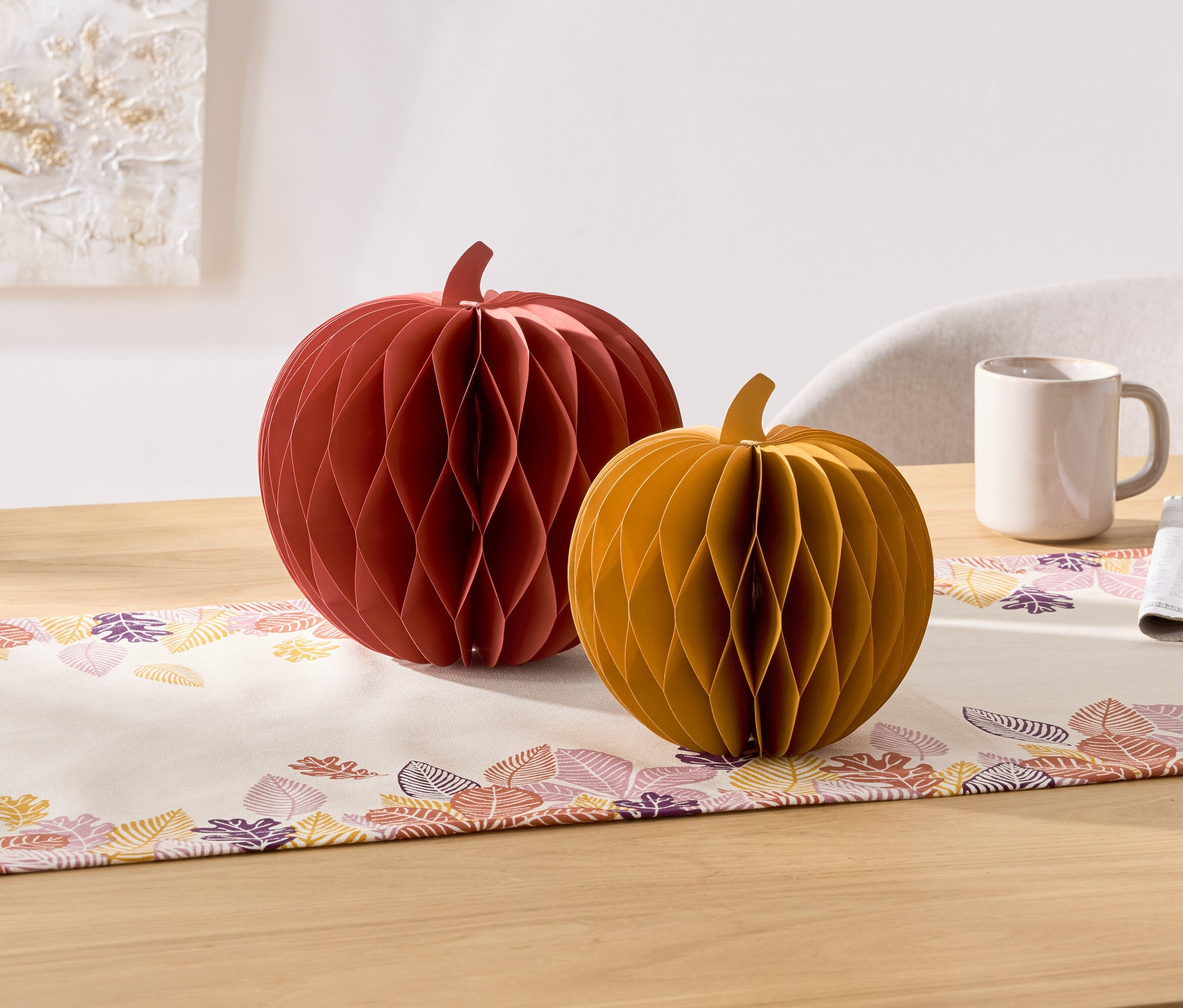 Na stole stoją dwie dekoracyjne dynie z papieru, czerwona i żółta. Obok nich znajduje się kubek i serwetka.