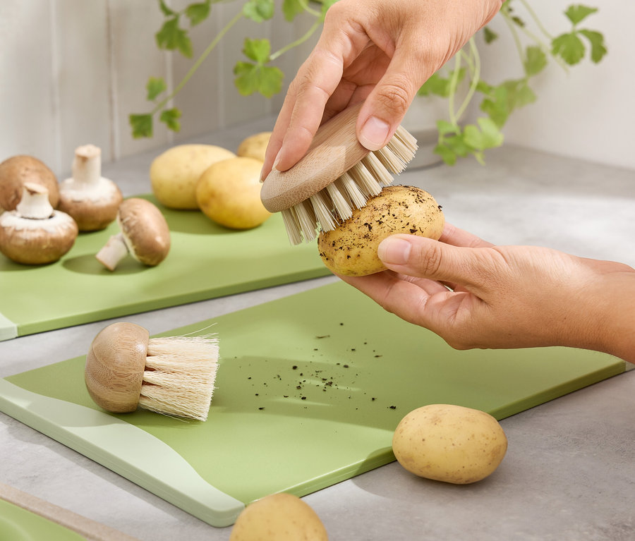 Ręce czyszczą szczotką ziemniaka na zielonej desce do krojenia. Na drugim planie brązowe pieczarki i ziemniaki.