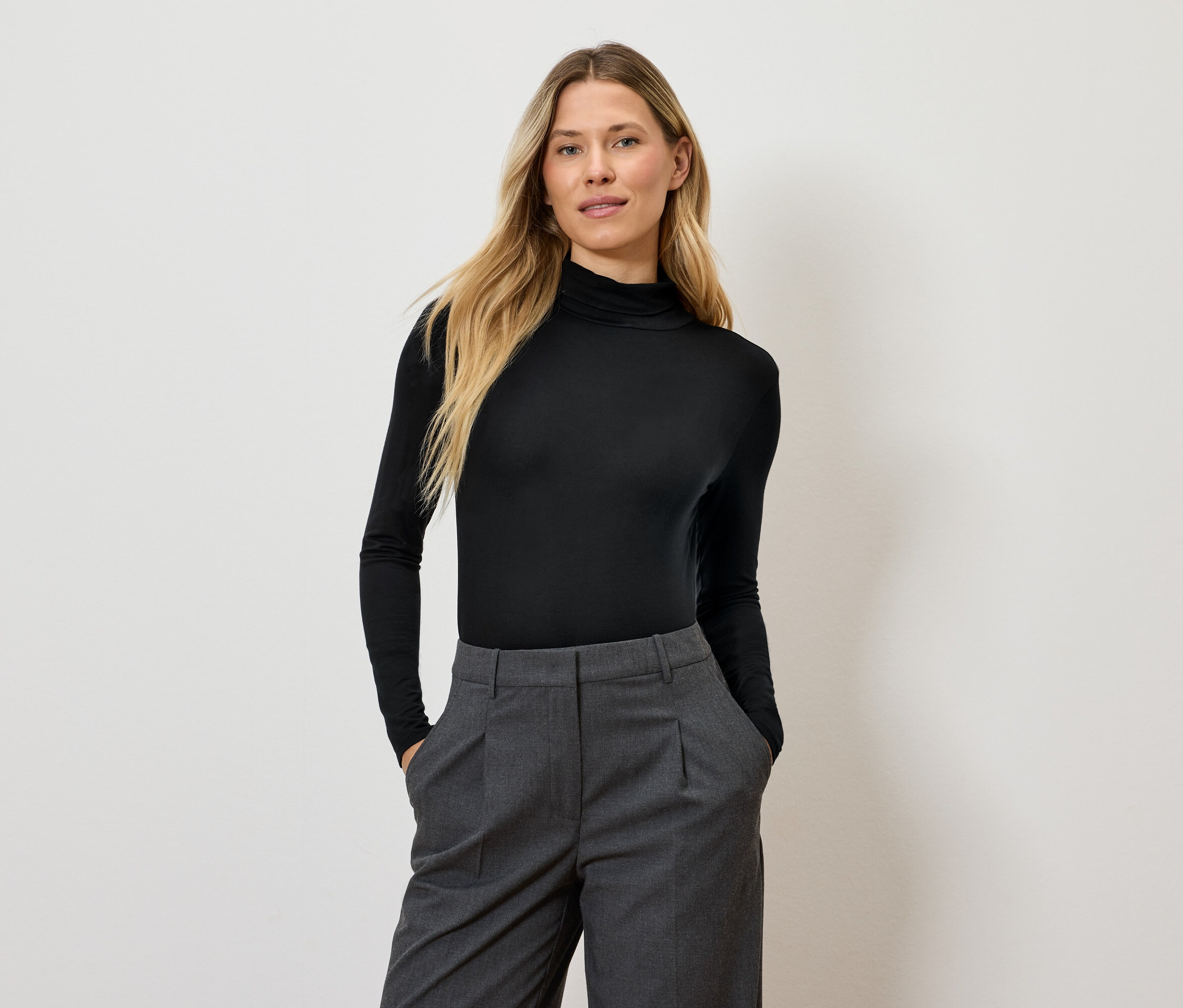 Kobieta z długimi blond włosami pozuje w czarnym golfie i szarych spodniach.