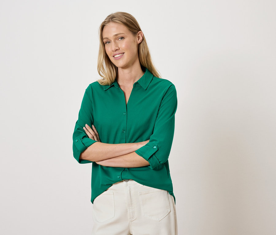 Kobieta pozuje ze skrzyżowanymi ramionami w zielonej koszuli i białych spodniach.