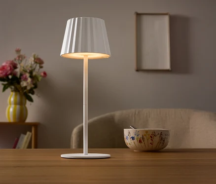 Na stole stoi biała lampa z plisowanym abażurem. W tle wazon z kwiatami i miska.
