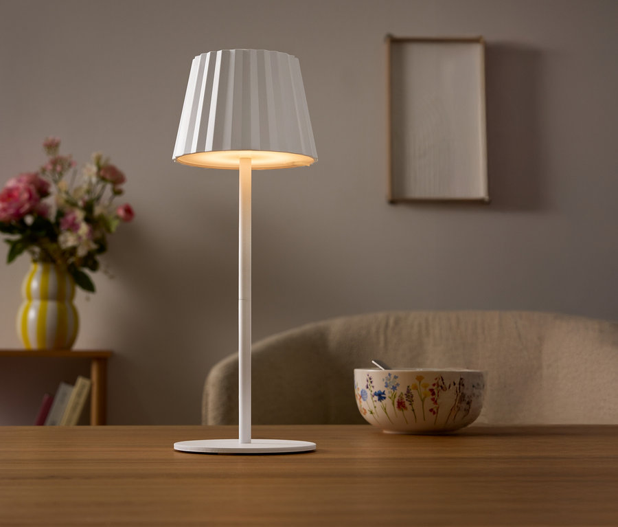 Na stole stoi biała lampa z plisowanym abażurem. W tle wazon z kwiatami i miska.