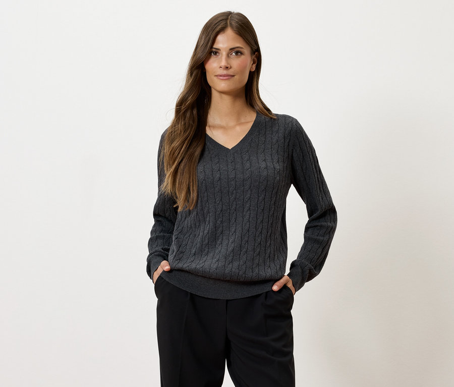 Kobieta z długimi brązowymi włosami pozuje na jasnym tle, ubrana w szary sweter z dzianiny w warkocze z dekoltem w serek i czarne spodnie.