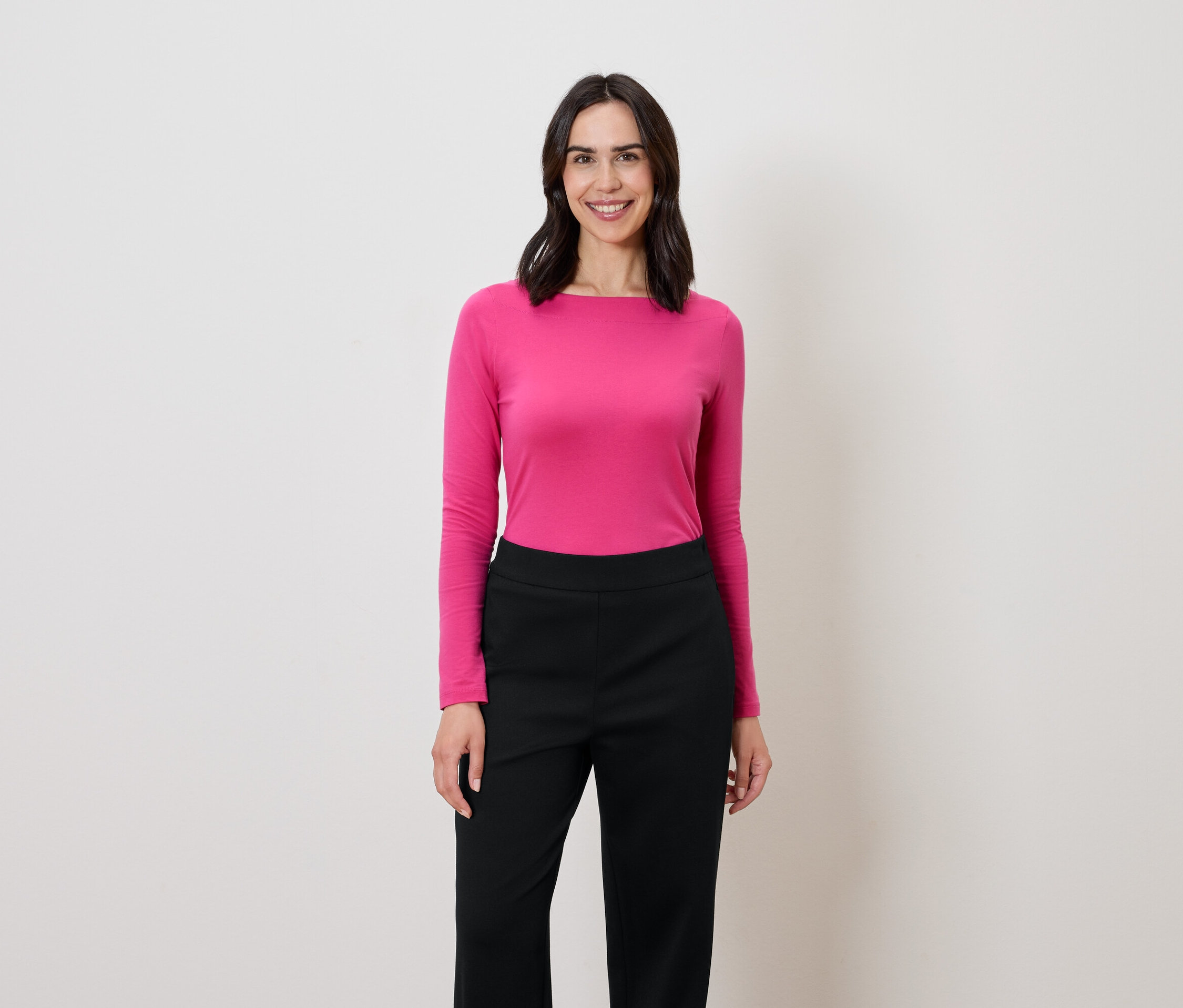 Kobieta o ciemnych włosach stoi i uśmiecha się, ma na sobie różową bluzkę z długim rękawem i czarne spodnie.