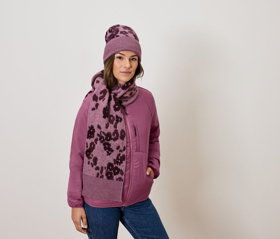 Kobieta ma na sobie różową kurtkę, dżinsy, szalik i czapkę w kwiaty.