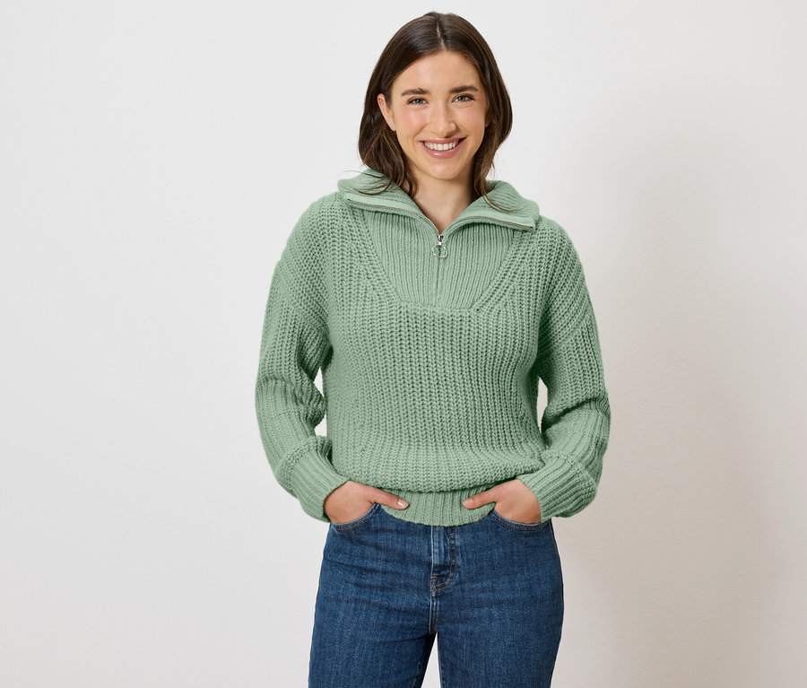Kobieta z brązowymi włosami pozuje w zielonym swetrze i niebieskich jeansach.