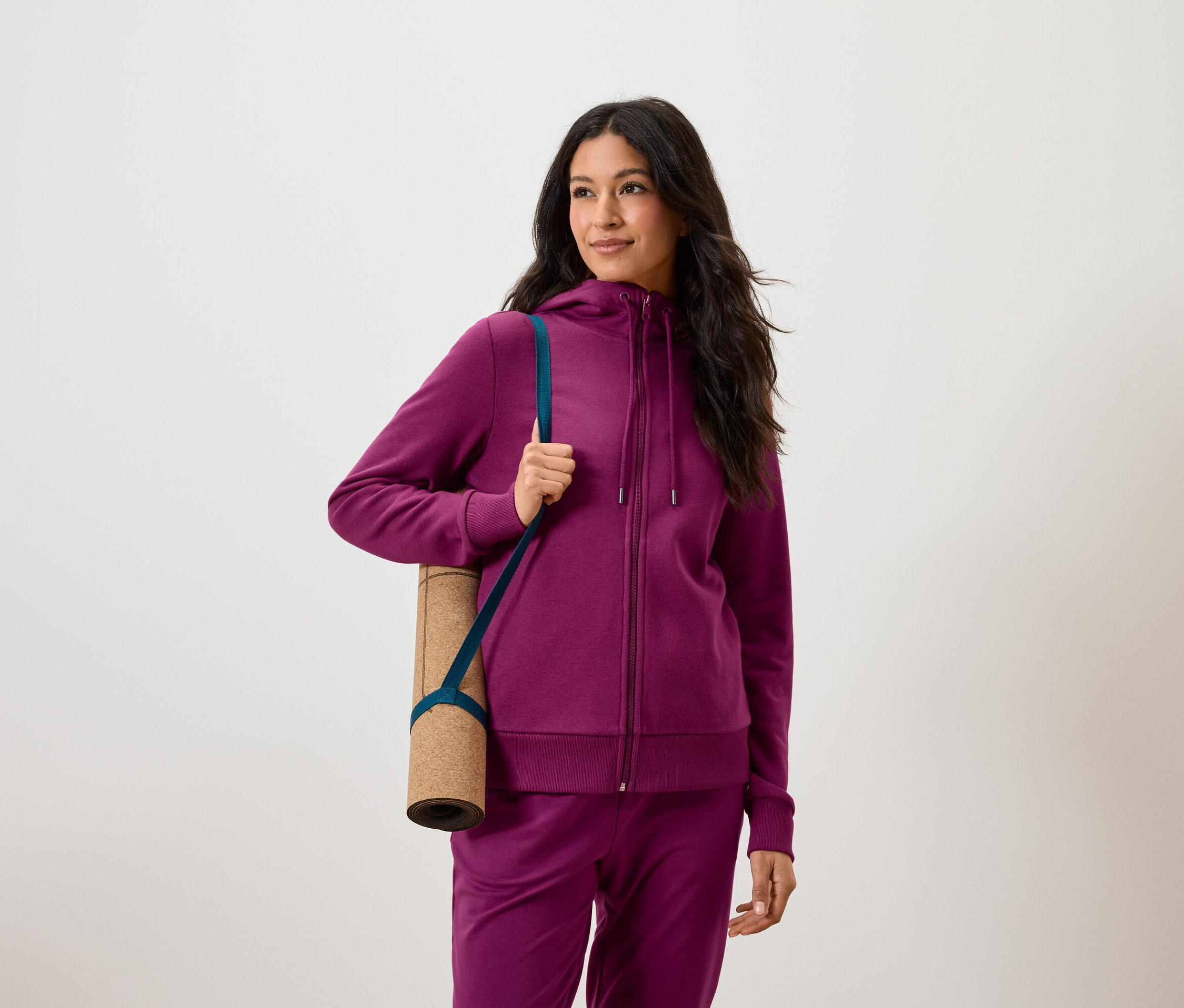 Kobieta z ciemnymi włosami w fioletowym dresie trzyma pod pachą zwiniętą matę do ćwiczeń.