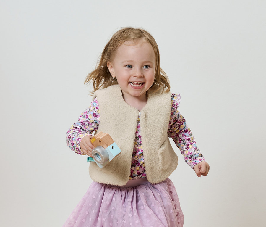 Uśmiechnięta dziewczynka z drewnianym aparatem-zabawką, ubrana w liliową bluzkę i spódnicę, na jasnym tle.