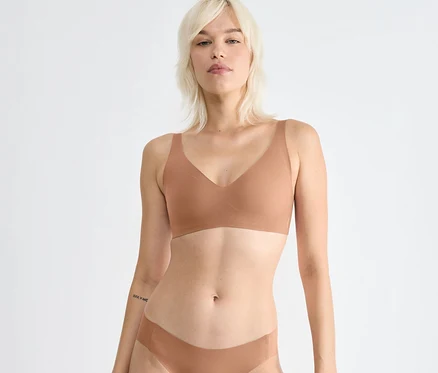 Blondynka w miękkim biustonoszu sloggi ZERO Feel 2.0 w kolorze nostalgic brown i pasującej bieliźnie, patrzy przed siebie. Ma tatuaż na lewym przedramieniu.