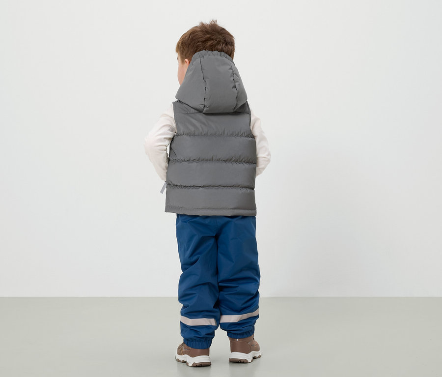 Chłopiec stoi tyłem, ubrany w szarą kamizelkę z kapturem, niebieskie spodnie i brązowe buty.