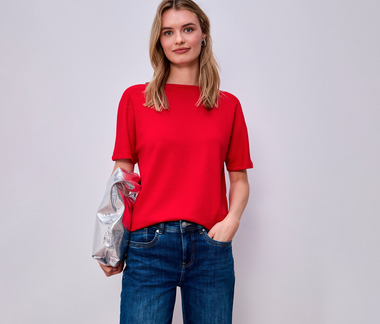 Kobieta w czerwonej koszulce Street One i dżinsach, srebrna kurtka na ramieniu.