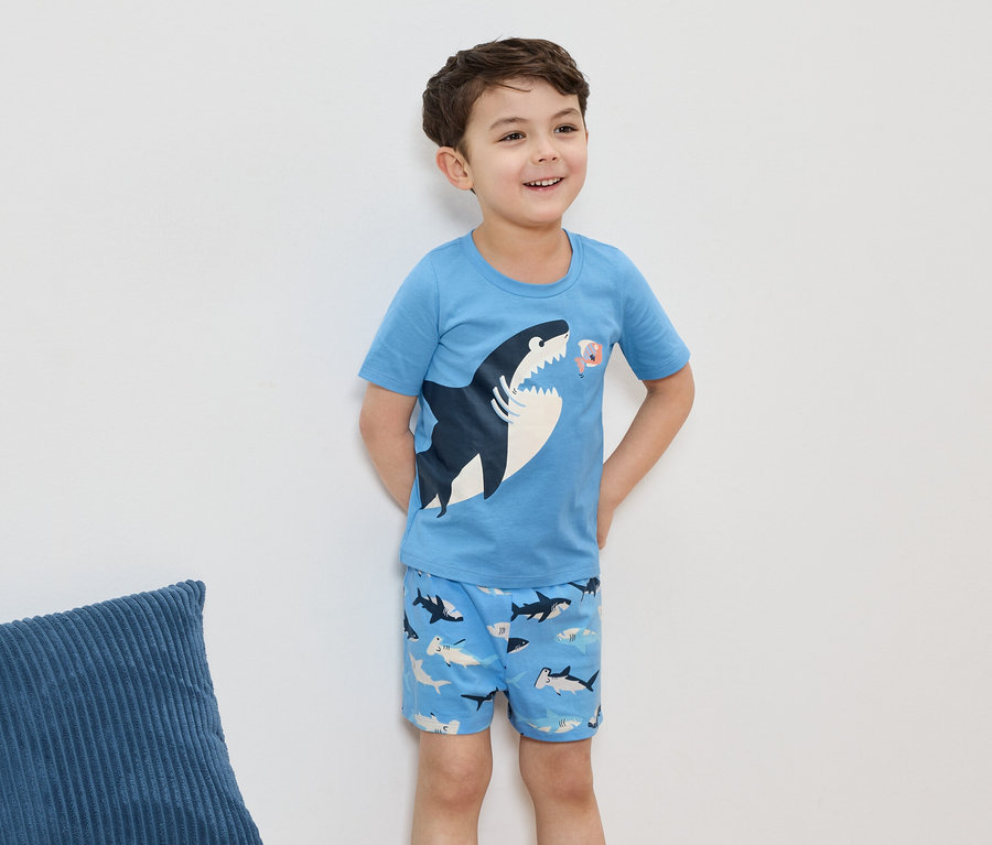 Uśmiechnięty chłopiec w jasnoniebieskiej piżamie z motywem rekina, na tle białej ściany.