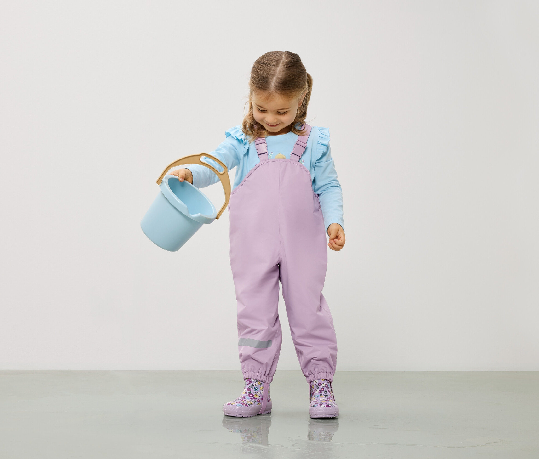 Dziewczynka w jasnoniebieskiej bluzce i liliowych ogrodniczkach trzyma jasnoniebieskie wiaderko.