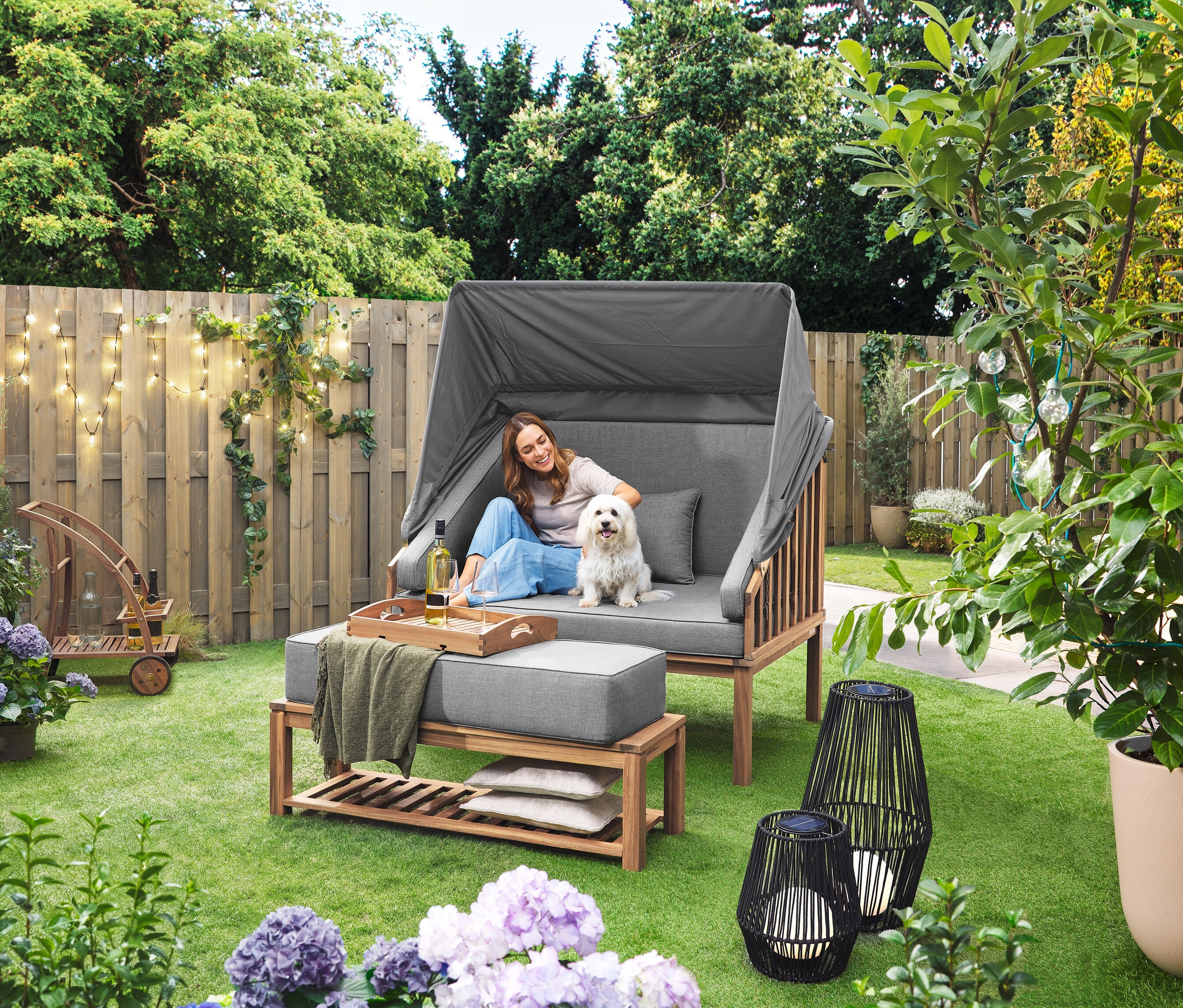 Kobieta siedzi z psem w koszu plażowym Premium Lounge-Strandkorb mit Sonnenschutz-Dach. Na trawniku stoi latarnia solarna ze sznurkiem tekstylnym, duża i latarnia solarna ze sznurkiem tekstylnym, mała. W tle widoczny jest wózek barowy z odłączaną tacą. Na płocie wisi łańcuch LED-Outdoor-Lichterkette mit Netzadapter.