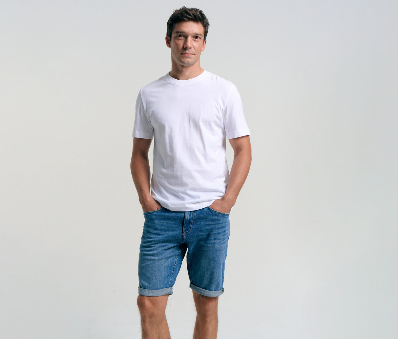 Mężczyzna w białym T-shircie i szortach dżinsowych Tom Tailor TTJOSH REGULAR SLIM, mid stone bright blue denim, z rękami w kieszeniach.