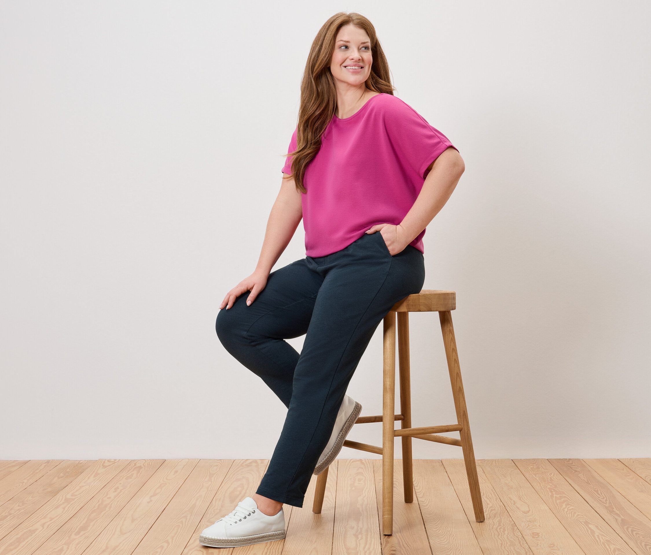 Kobieta siedzi na stołku, ma na sobie różową bluzkę, niebieskie spodnie i białe buty.