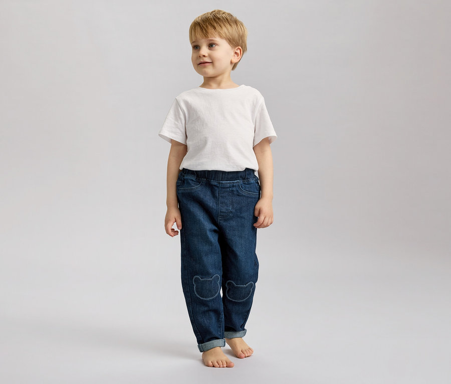 Chłopiec o blond włosach stoi boso na szarym tle, ubrany w dżinsy z motywem misia i biały T-shirt.