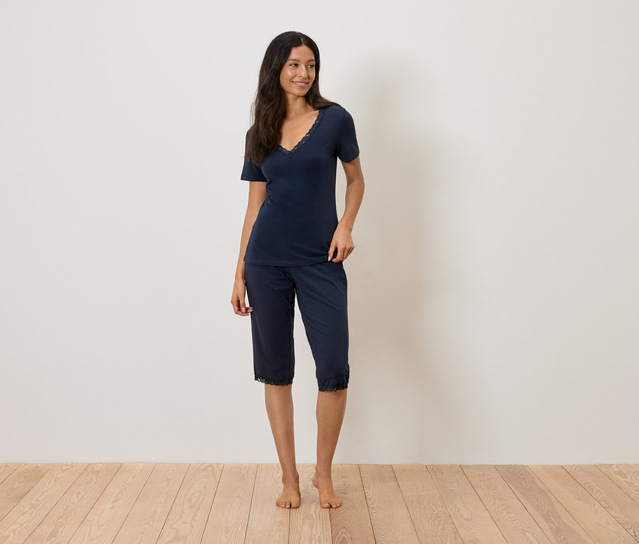 Kobieta stoi boso na drewnianej podłodze, ubrana w granatową piżamę z koronkowym wykończeniem.