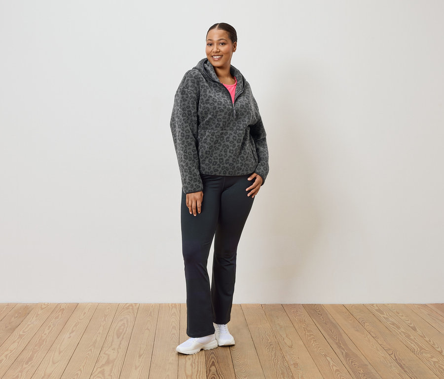 Kobieta stoi i ma na sobie szaro-czarną bluzę z kapturem z polaru w panterkę, czarne spodnie i białe tenisówki.