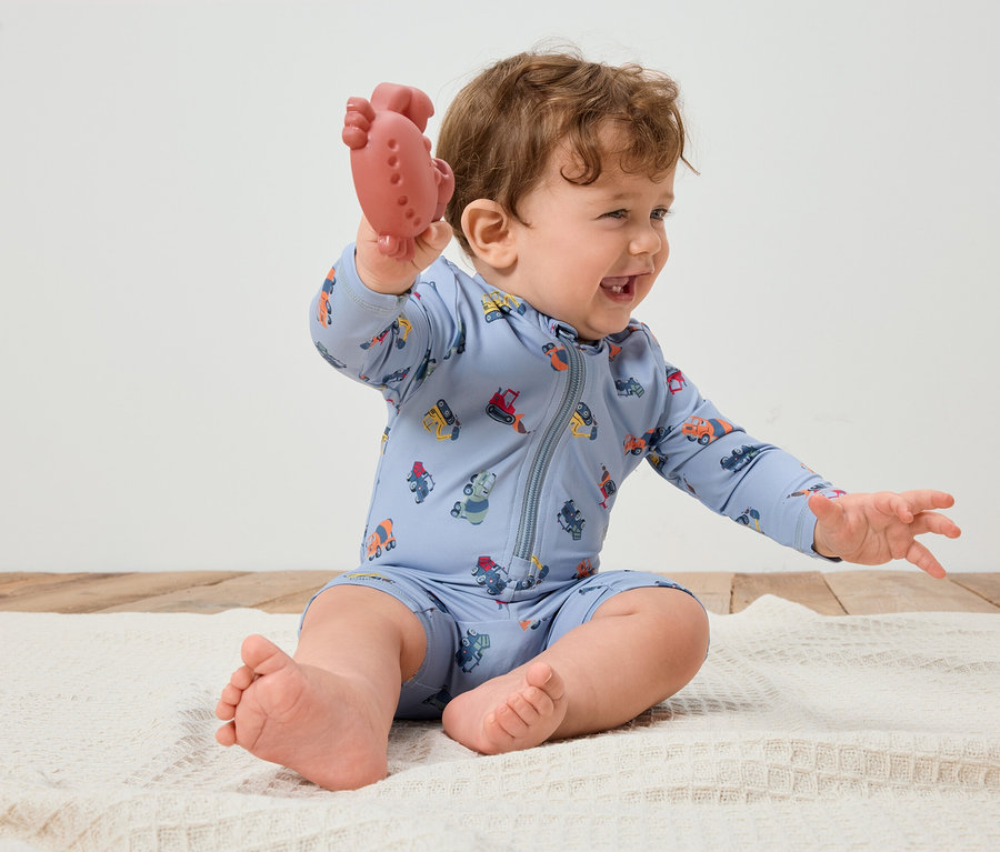 Uśmiechnięty niemowlak z kręconymi włosami siedzi na białym kocyku, trzymając w ręku czerwony gryzak. Niemowlak ubrany jest w jasnoniebieski kombinezon z kolorowymi pojazdami.