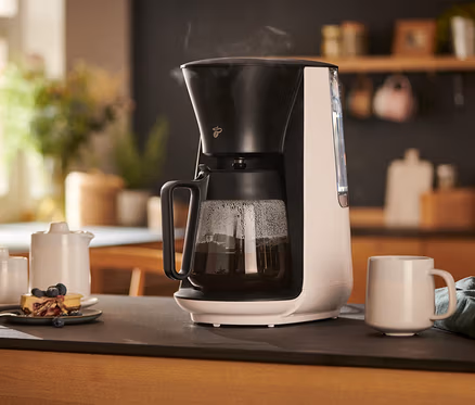 Biały ekspres przelewowy »Let's Brew« stoi na stole obok filiżanki, dzbanka na mleko, cukierniczki i talerza z ciastem.