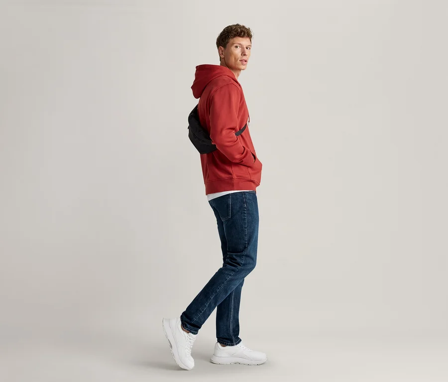 Mężczyzna w czerwonej bluzie z kapturem, niebieskich dżinsach i białych sneakersach, z czarną nerką przewieszoną przez klatkę piersiową, ręce w kieszeniach, patrzy przez ramię.