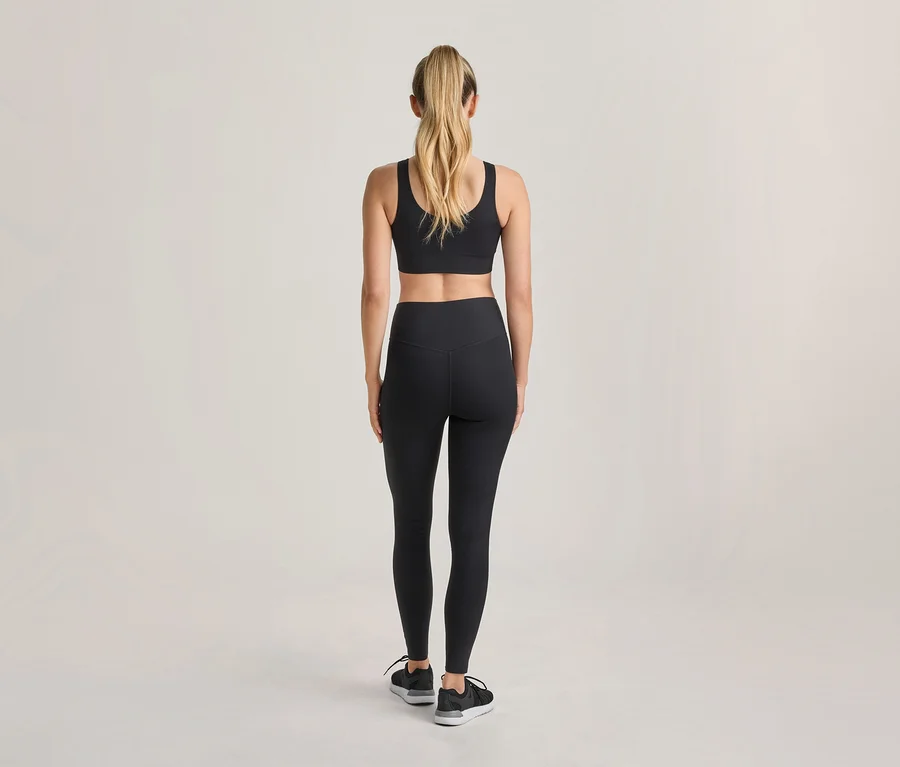 Kobieta widziana od tyłu, ubrana w czarny biustonosz sportowy, czarne legginsy i czarne buty sportowe, z blond włosami spiętymi w kucyk.