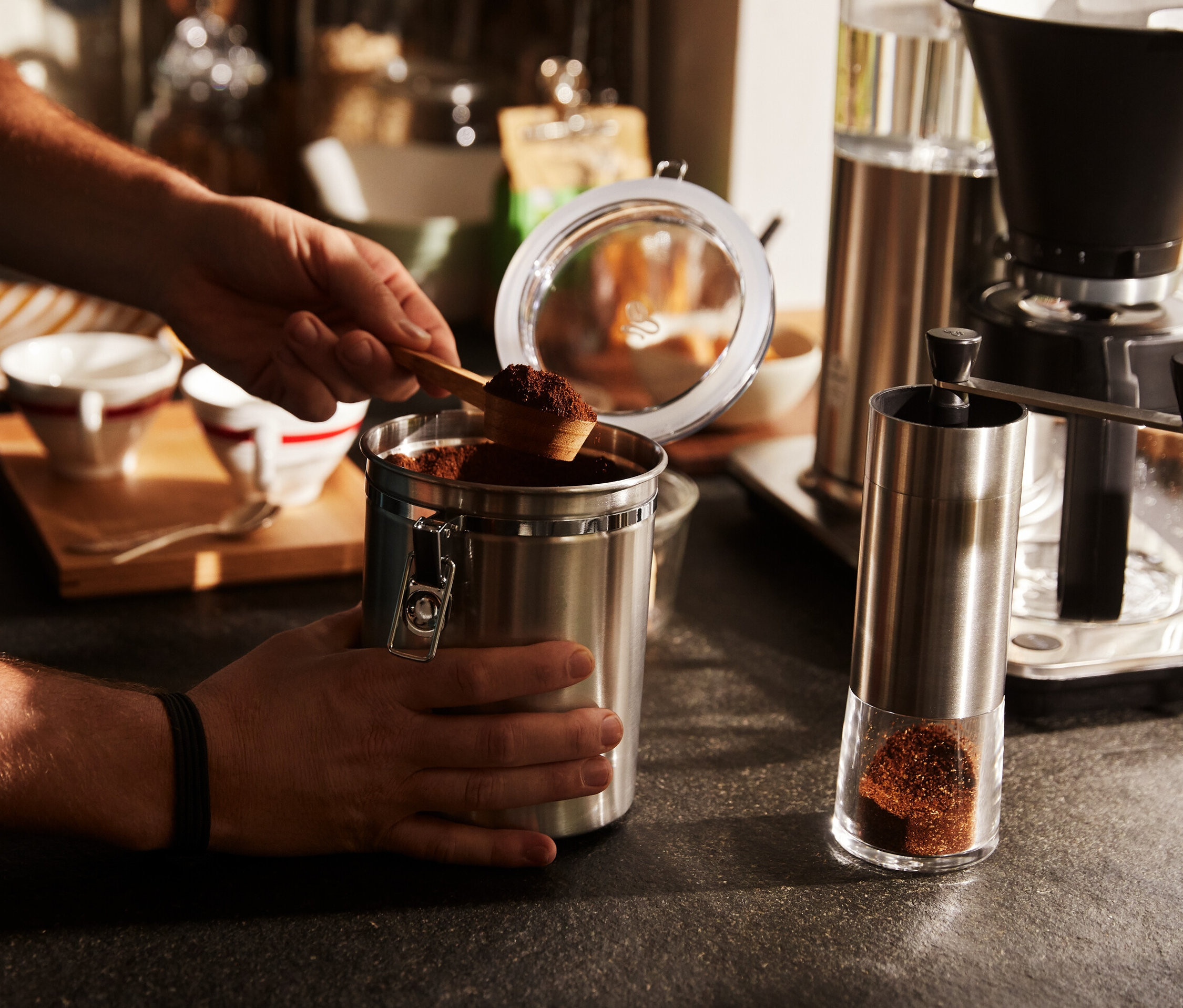 Mężczyzna nabiera łyżką kawę z metalowej puszki. Obok stoi srebrny ręczny młynek do kawy.