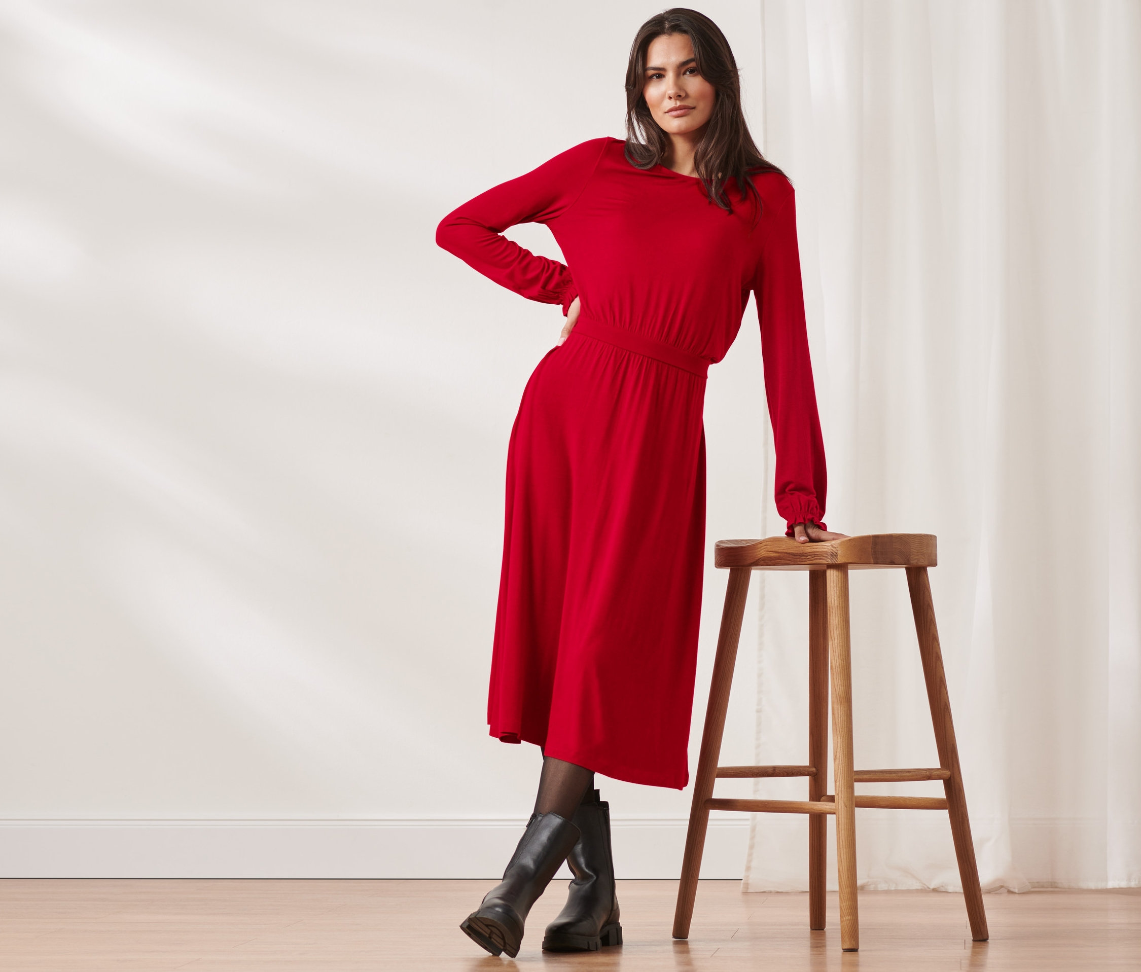 Kobieta pozuje w czerwonej sukience dżersejowej o długości midi i czarnych butach przy drewnianym stołku.