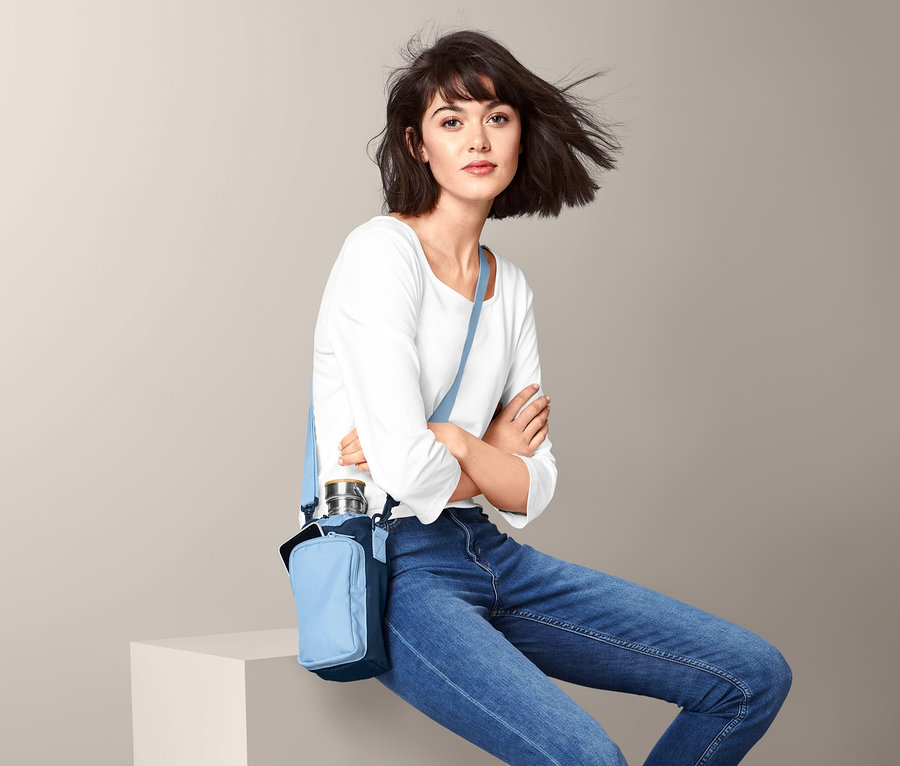 Kobieta siedzi ze skrzyżowanymi ramionami. Nosi torbę na ramię na butelkę.