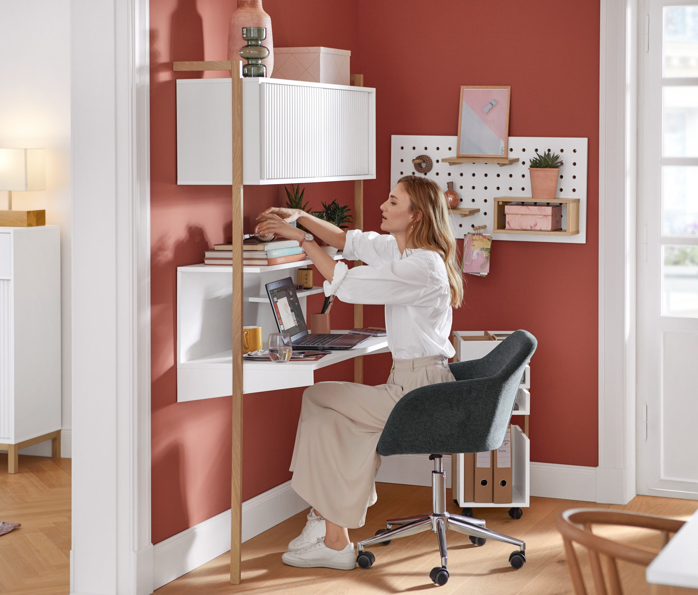 Kobieta siedzi na beżowym krześle do biurka przy biurku z regałem i pracuje na laptopie.