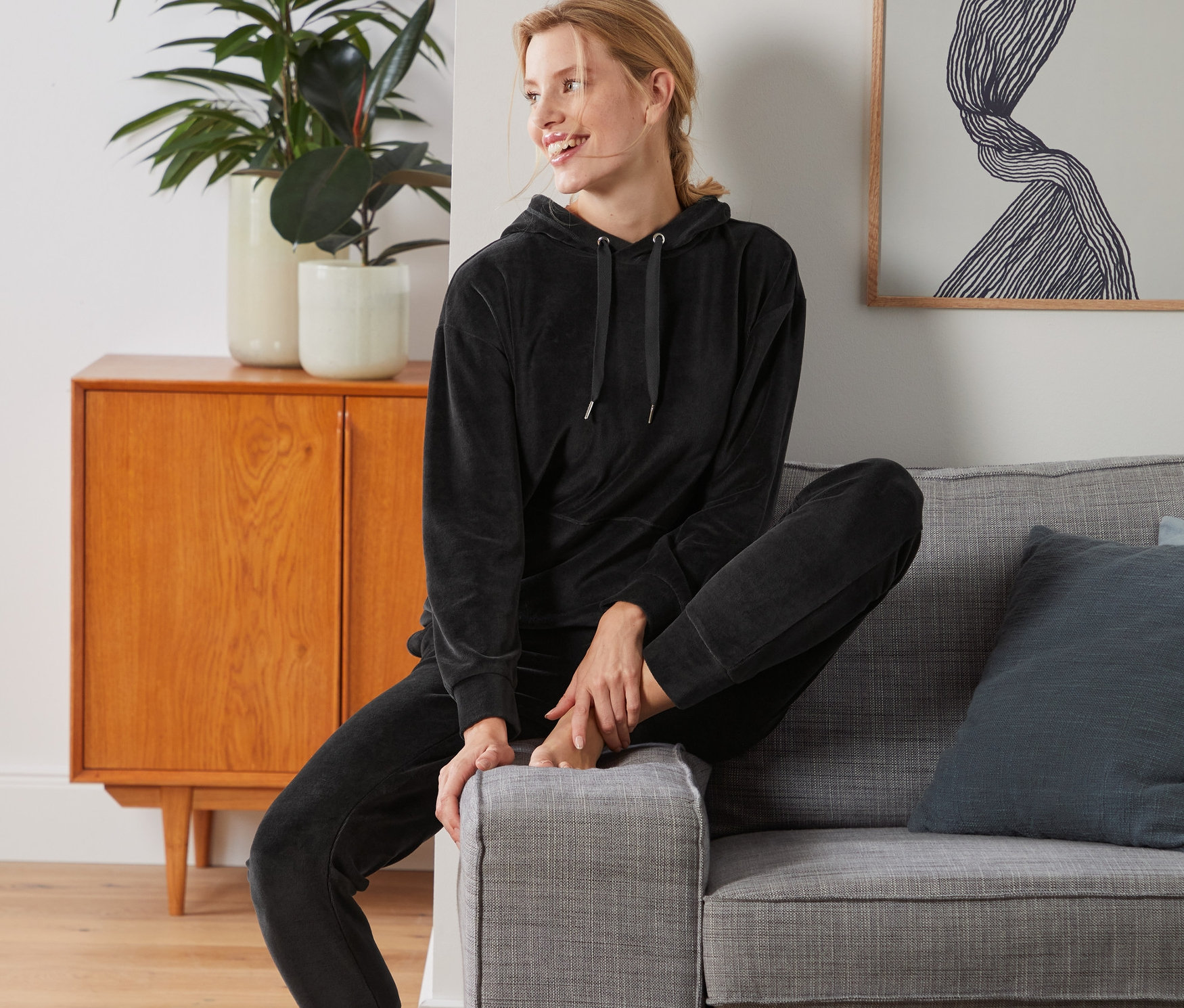 Kobieta siedzi na szarej sofie, ubrana w antracytowy dres domowy z weluru.