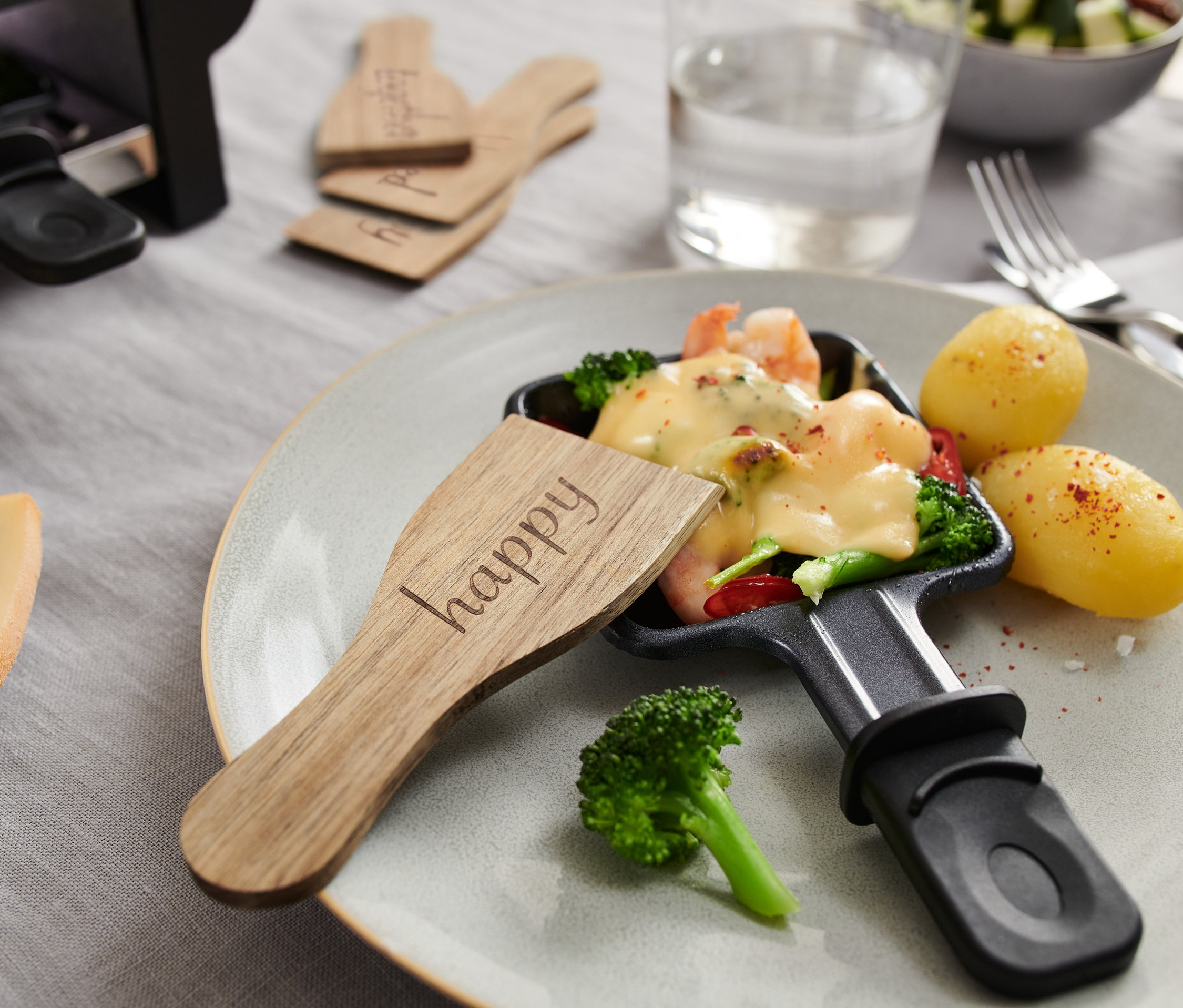 Zbliżenie na patelnię do raclette z roztopionym serem, warzywami i ziemniakami, z drewnianymi Suwaki do raclette, 4 sztuki z napisem "happy".
