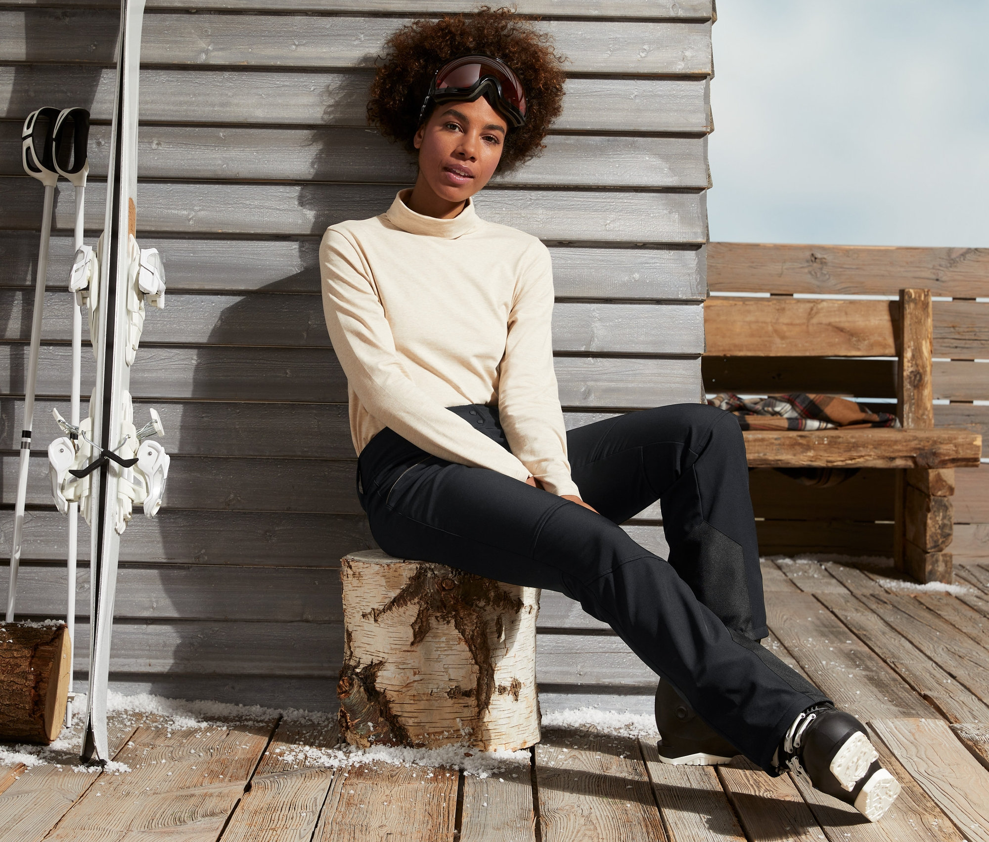 Kobieta siedzi na drewnianym klocu w czarnych spodniach narciarskich i butach narciarskich.