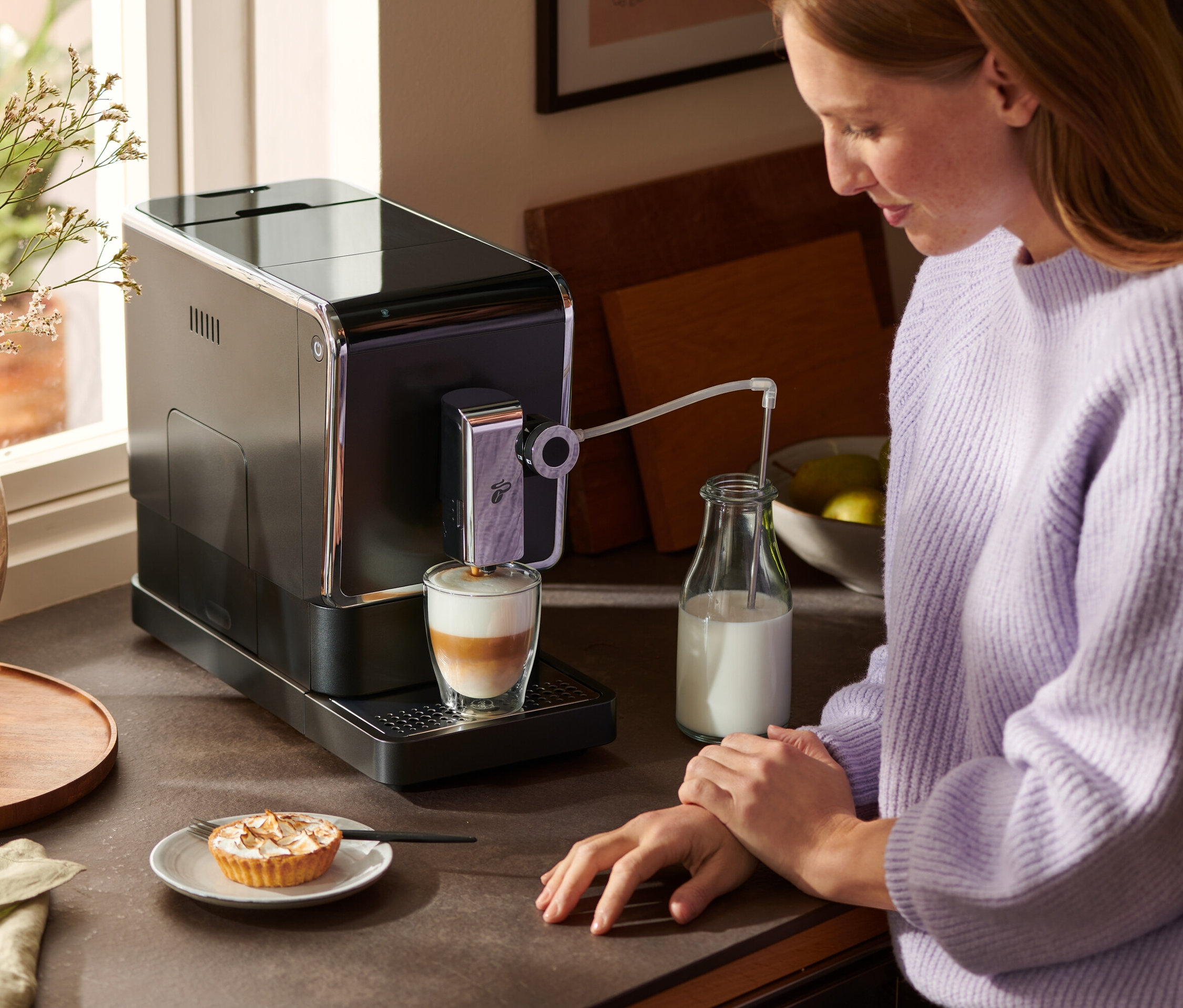 Kobieta stoi przy blacie i używa automatycznego ekspresu do kawy Tchibo »Esperto Pro« w kolorze antracytowym do przygotowania latte. Obok ekspresu stoi butelka mleka i talerz ciasta.