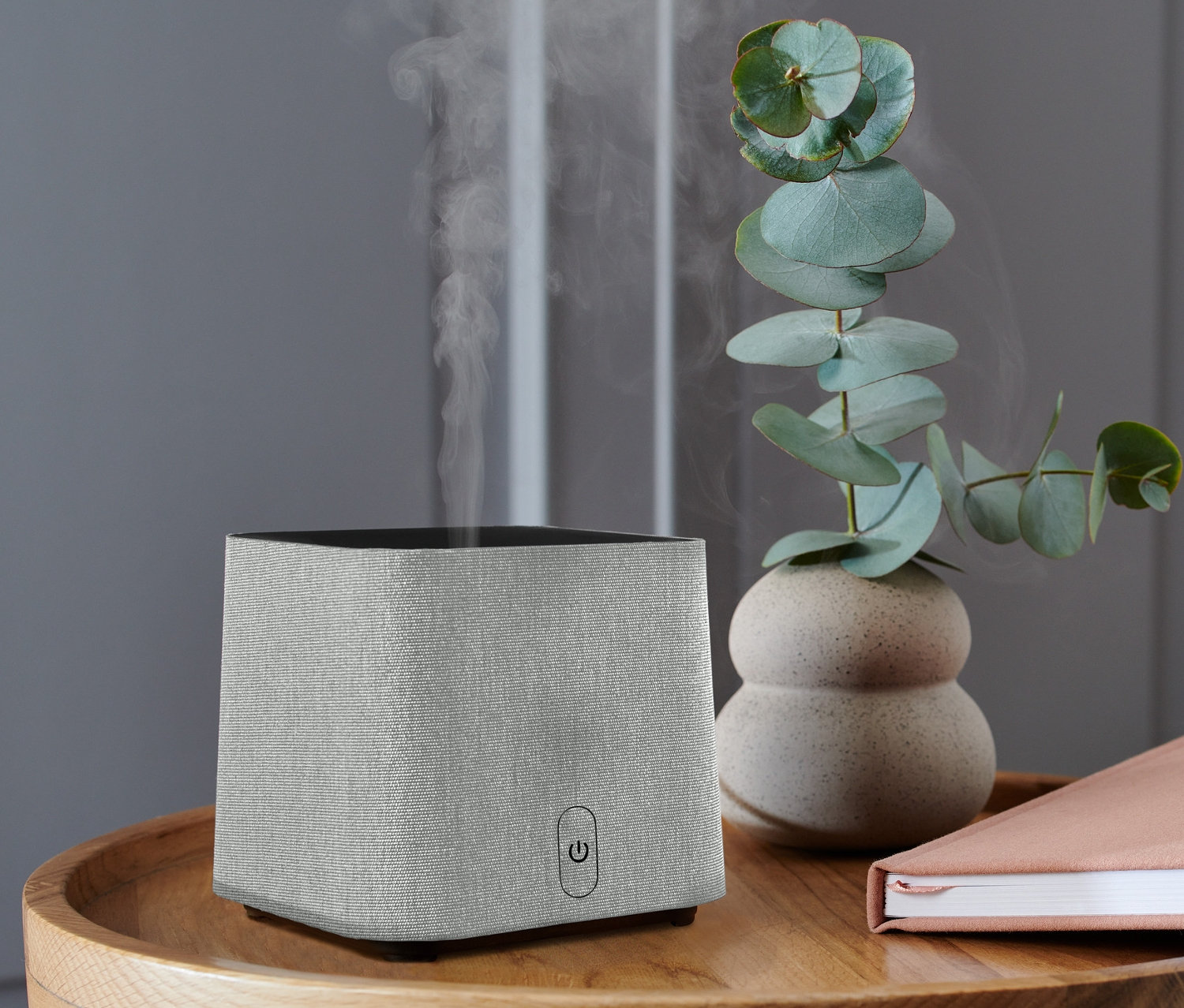 Szary dyfuzor zapachowy stoi na drewnianym stoliku z książką i wazonem eukaliptusa.