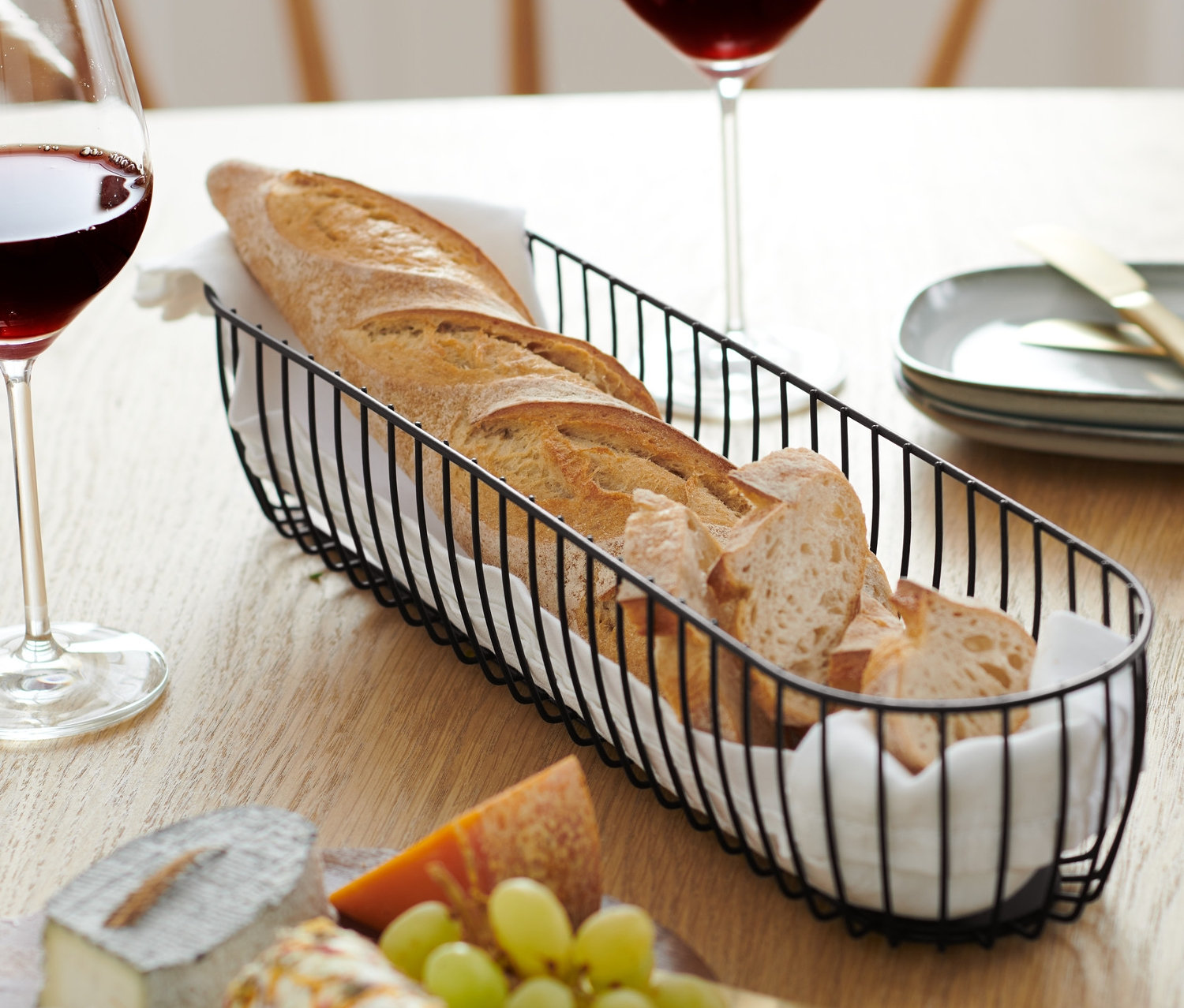 Na drewnianym stole stoją dwa kieliszki czerwonego wina, miska druciana z bagietką i deska serów z winogronami.