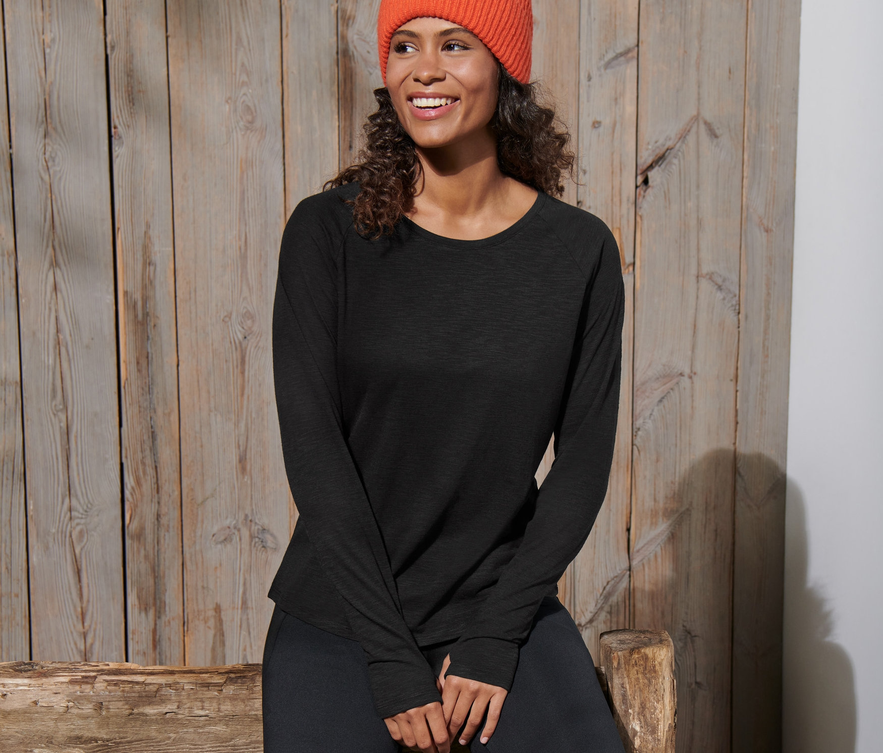 Kobieta siedzi na drewnianej ławce, ubrana w czarną koszulkę sportową z długim rękawem i pomarańczową czapkę.