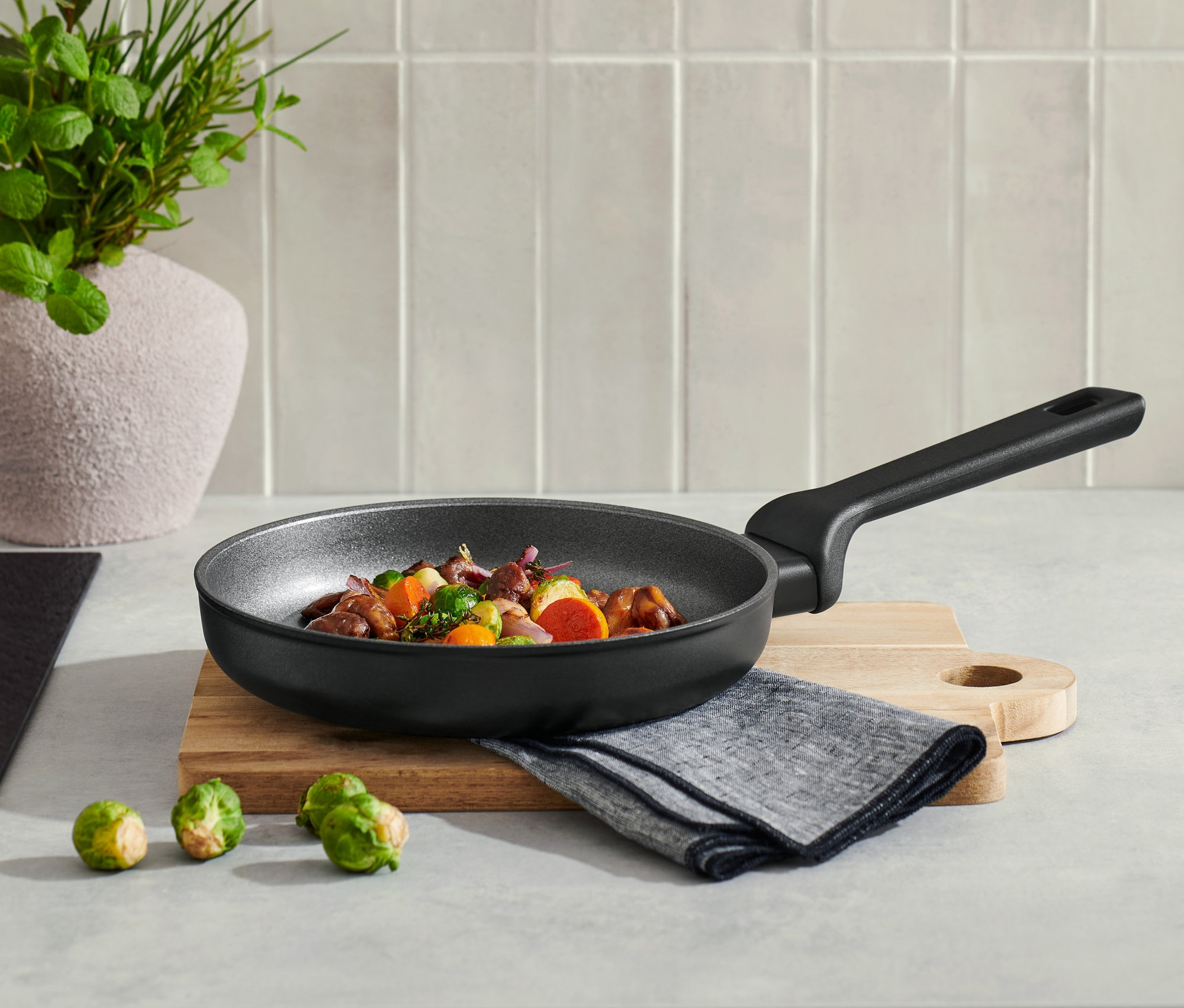 Na blacie kuchennym stoi profesjonalna patelnia aluminiowa o średnicy ok. 20 cm z zawartością, obok leżą trzy brukselki.