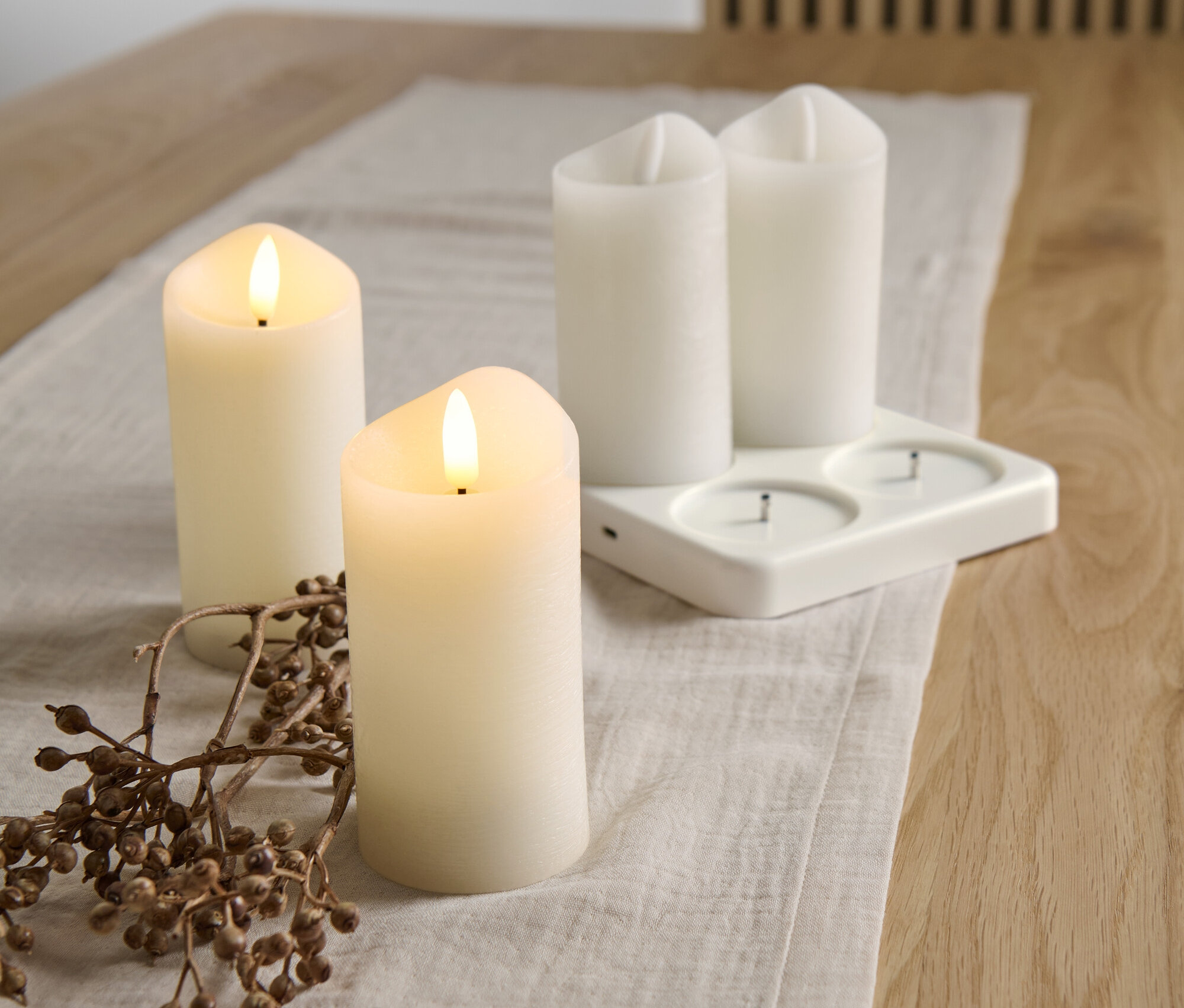 Na stole leżą świece z prawdziwego wosku LED z zasilaniem akumulatorowym, 4 sztuki, ze stacją ładującą i gałązka z jagodami.