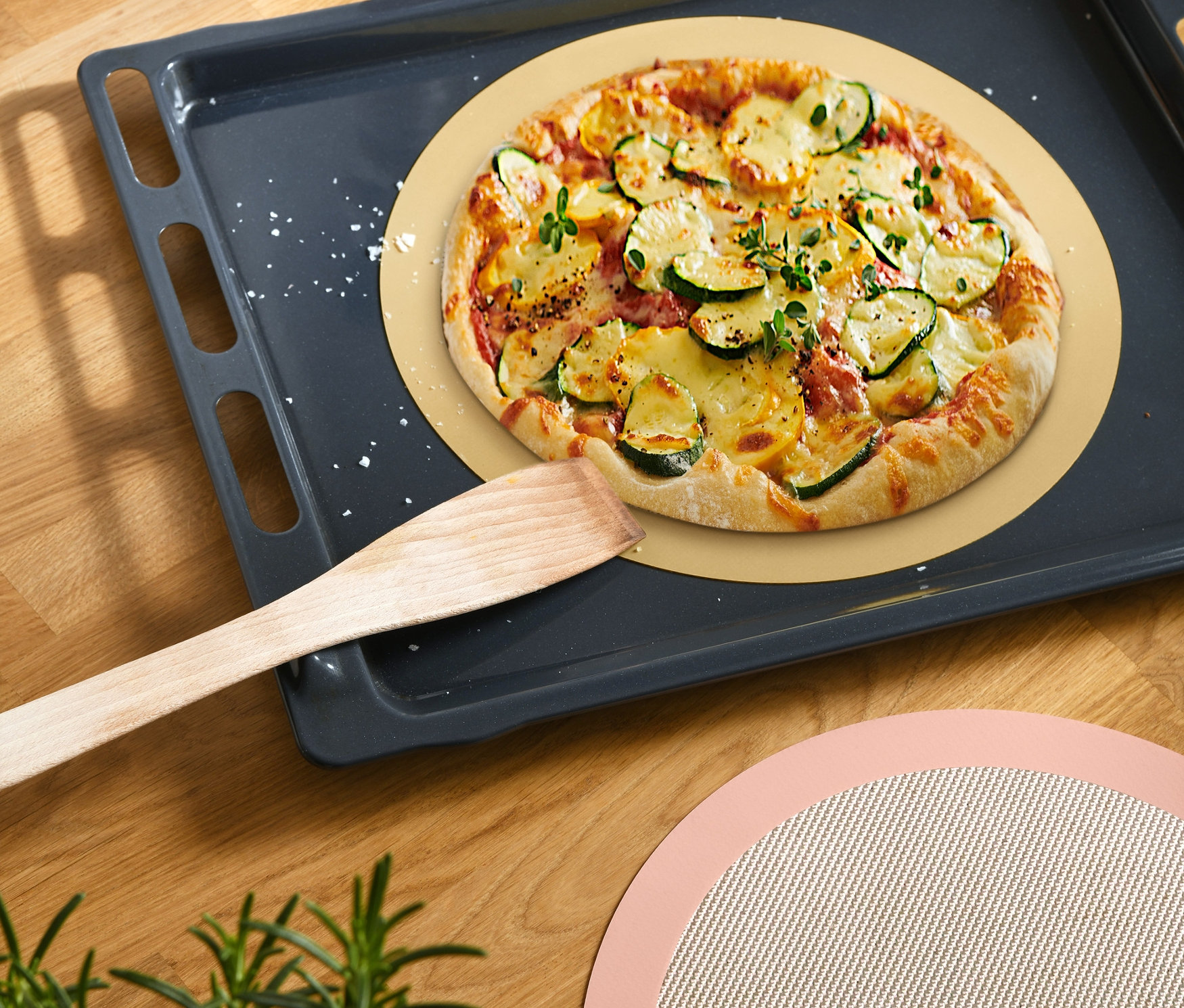 Pizza na okrągłej macie do pieczenia na blasze do pieczenia z drewnianą szpatułką obok i drugą okrągłą matą do pieczenia obok.