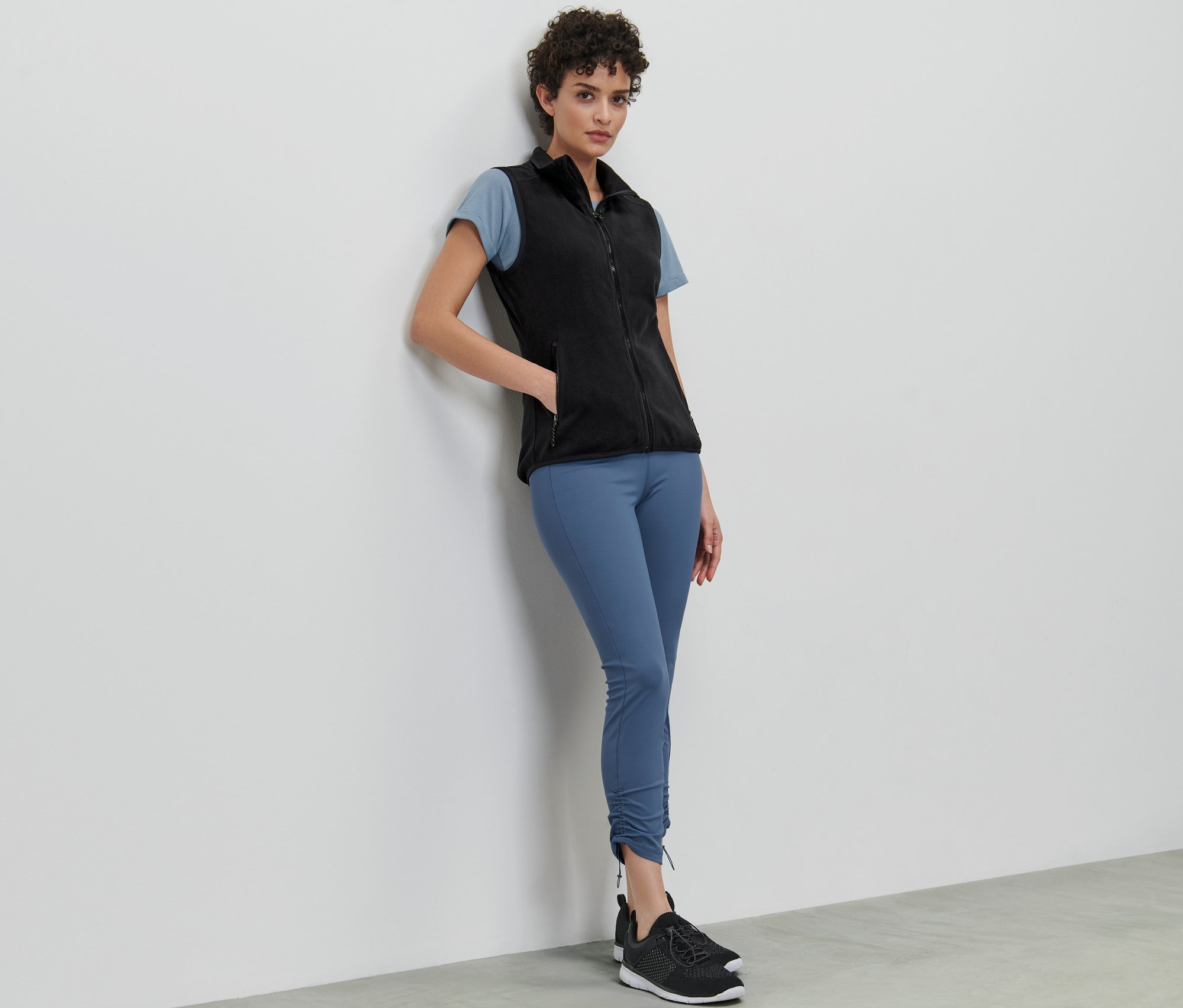 Kobieta oparta o ścianę ma na sobie kamizelkę z polaru, t-shirt i legginsy.
