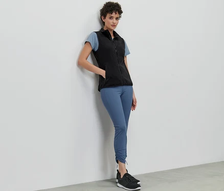 Kobieta oparta o ścianę ma na sobie kamizelkę z polaru, t-shirt i legginsy.